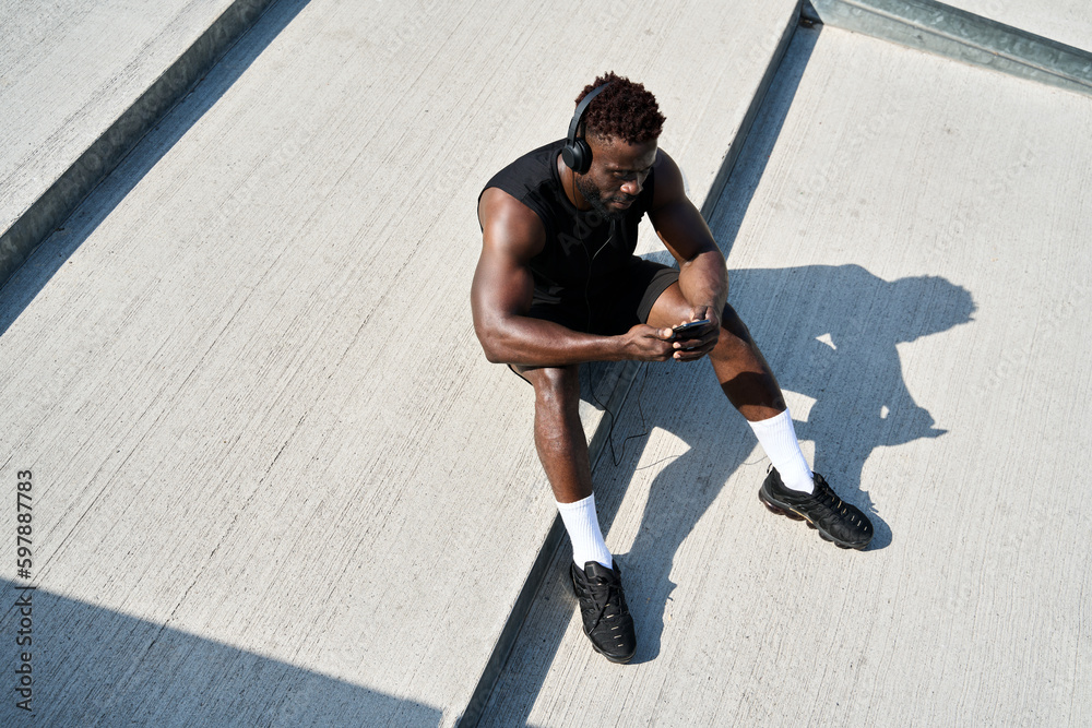 Fit sporty young black man sitting on concrete urban stairs holding phone using mobile apps listening music. Strong African ethnic guy wearing headphones looking at smartphone outdoors. Top view - obrazy, fototapety, plakaty 