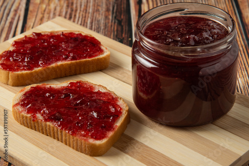 Delicious toasts with sweet jams on marble background, Raspberry jam set. Confiture spread on piece of toast bread, knife, glass jar with jelly, spoon, bowl and fresh red strawberry