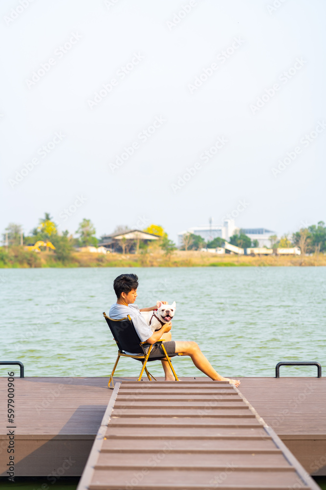 Asian man playing with his French bulldog during resting by the lake at pets friendly park. Domestic dog with owner enjoy outdoor lifestyle travel nature on summer vacation. Pet Humanization concept.