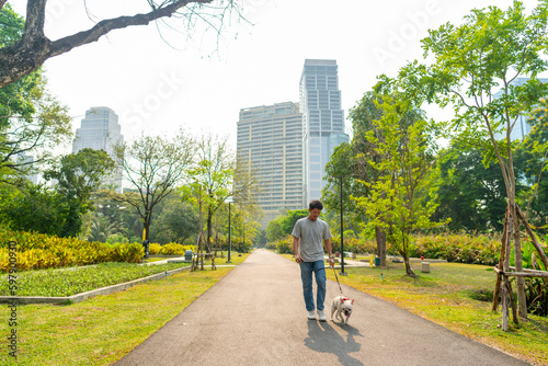 Asian man walking with french bulldog breed at pets friendly dog park. Domestic dog with owner enjoy urban outdoor lifestyle in the city on summer vacation. Pet Humanization and pet parents concept.