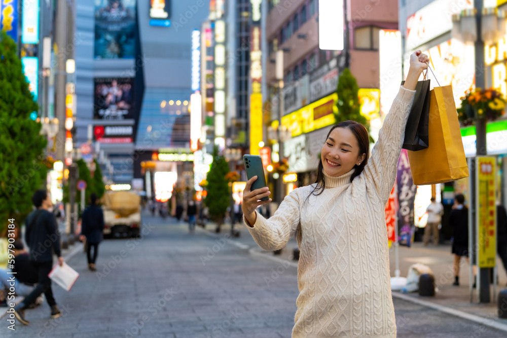 Fototapeta premium Happy Asian woman using mobile phone taking selfie during shopping at Tokyo city, Japan. Attractive girl enjoy outdoor lifestyle travel city street with using wireless technology on holiday vacation.