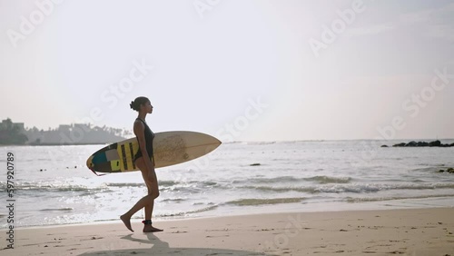 African american woman walking with surfboard on ocean beach. Black female surfer going with surf board. Pretty multiethnic girl goes on surfing session at sunrise. Slow motion, side view.