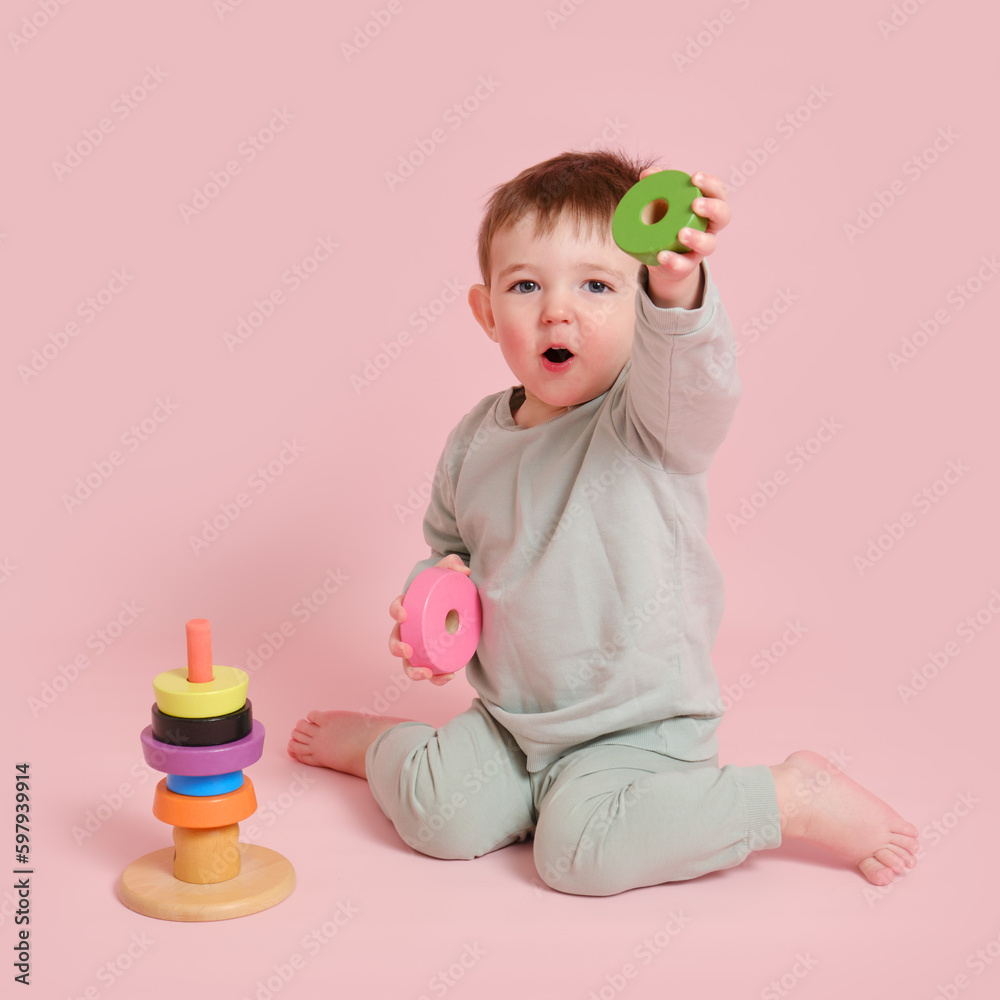 Happy toddler baby plays with a pyramid on a studio pink background. Child boy puts wooden rings on a toy pyramid. Kid age one year eight months, full height Happy toddler baby plays with a pyramid on a studio pink background. Child boy puts wooden rings on a toy pyramid. Kid age one year eight months, full height