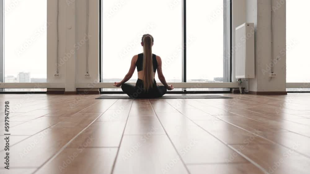 Rear view of young light-haired female in tight sports clothes sitting in lotus pose on yoga mat in front of window in fitness studio. Relaxed yogini practising Padmasana for deep meditation. Rear view of young light-haired female in tight sports clothes sitting in lotus pose on yoga mat in front of window in fitness studio. Relaxed yogini practising Padmasana for deep meditation.