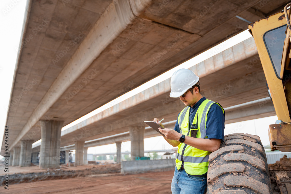 A civil engineer logging data with ipad standing beside industrial grader wheels at road construction site.