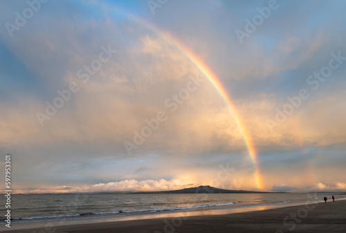 Rainbows over Rangitoto Island. Unrecognizable people walking on Milford beach. Auckland.