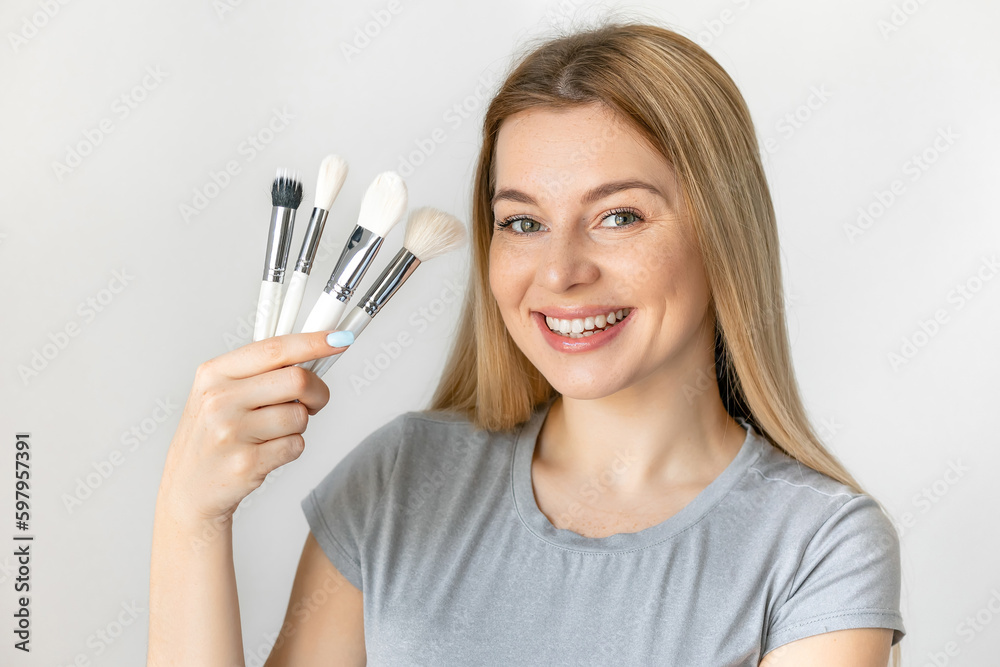 Portrait of beautiful cheerful make up with long hair with variety of brushes in arm, looking at camera, standing over white background Portrait of beautiful cheerful make up with long hair with variety of brushes in arm, looking at camera, standing over white background