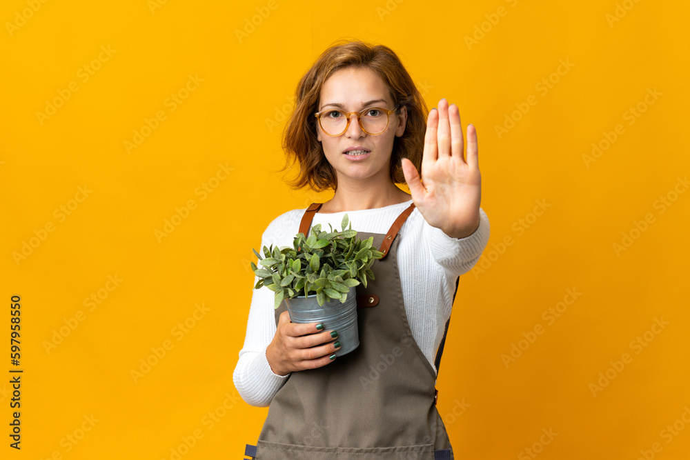 Young Georgian woman holding a plant isolated on yellow background making stop gesture Young Georgian woman holding a plant isolated on yellow background making stop gesture