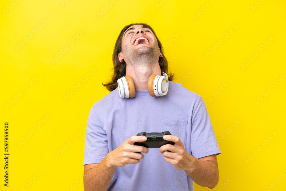 Young handsome caucasian man playing with a video game controller over isolated on yellow background laughing Young handsome caucasian man playing with a video game controller over isolated on yellow background laughing