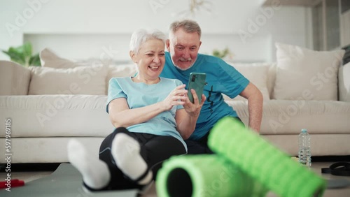 Laughing happy senior couple watch funny video on smartphone sit on couch after sport training in living roon at home. Browsing internet, social media watching, use online technologies aged people. photo