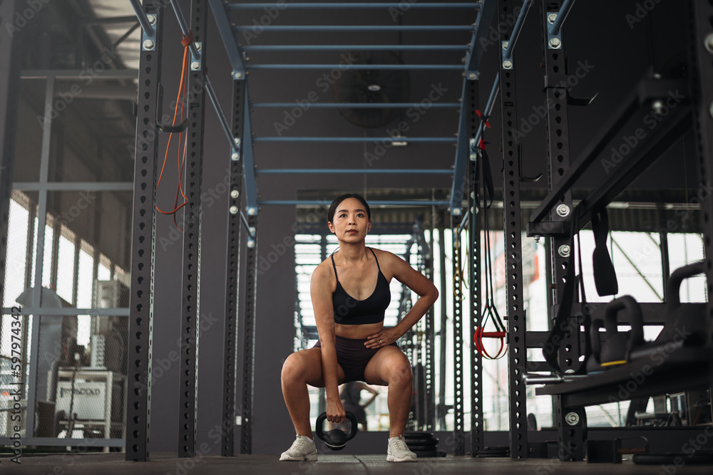 Strong asian woman doing exercise with kettlebell at crossfit gym. Athlete female wearing sportswear workout on grey gym background with weight and dumbbell equipment. Healthy lifestyle. Strong asian woman doing exercise with kettlebell at crossfit gym. Athlete female wearing sportswear workout on grey gym background with weight and dumbbell equipment. Healthy lifestyle.