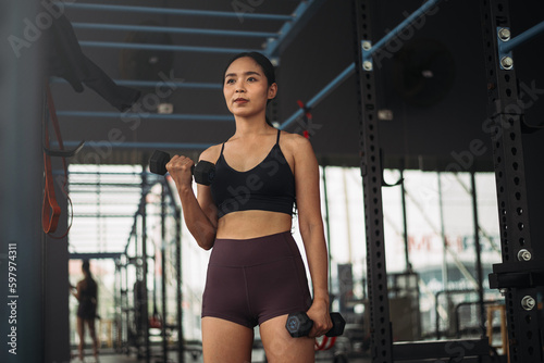 Strong Asian woman doing exercise with dumbbell at gym. Athlete female wearing sportswear workout on grey gym background with weight and dumbbell equipment. Healthy lifestyle.