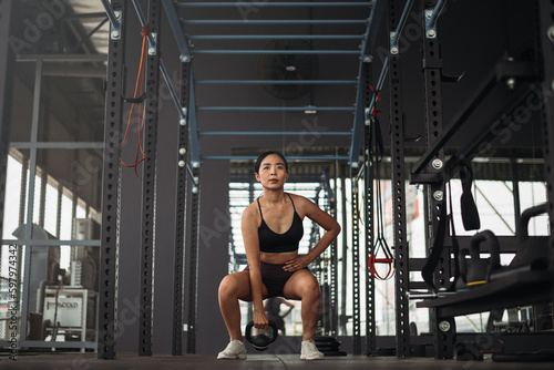 Strong asian woman doing exercise with kettlebell at crossfit gym. Athlete female wearing sportswear workout on grey gym background with weight and dumbbell equipment. Healthy lifestyle.