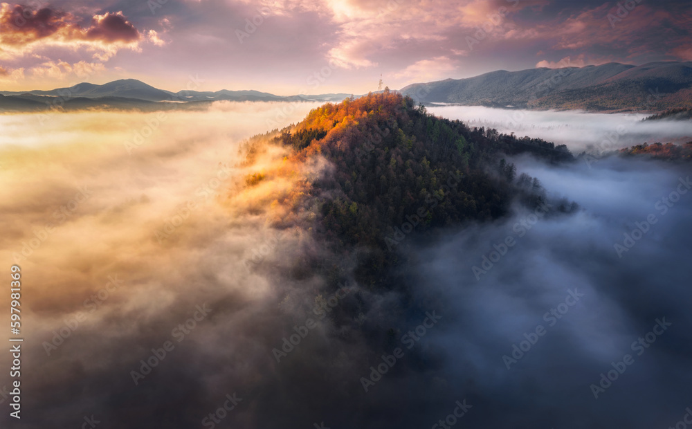 Aerial view of Mountain peak with sunlight and colorful forest in fog. Drone view on the beautiful colorful autumn mountains hills in low clouds at sunset or sunrise. Slovenia