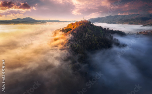 Aerial view of Mountain peak with sunlight and colorful forest in fog. Drone view on the beautiful colorful autumn mountains hills in low clouds at sunset or sunrise. Slovenia