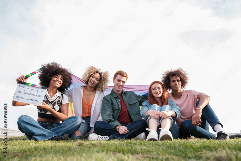 Obraz premium Group of heterogeneous young people sitting on the grass in the park with lgtbi flag. Concept: Pride, lifestyle, friendship