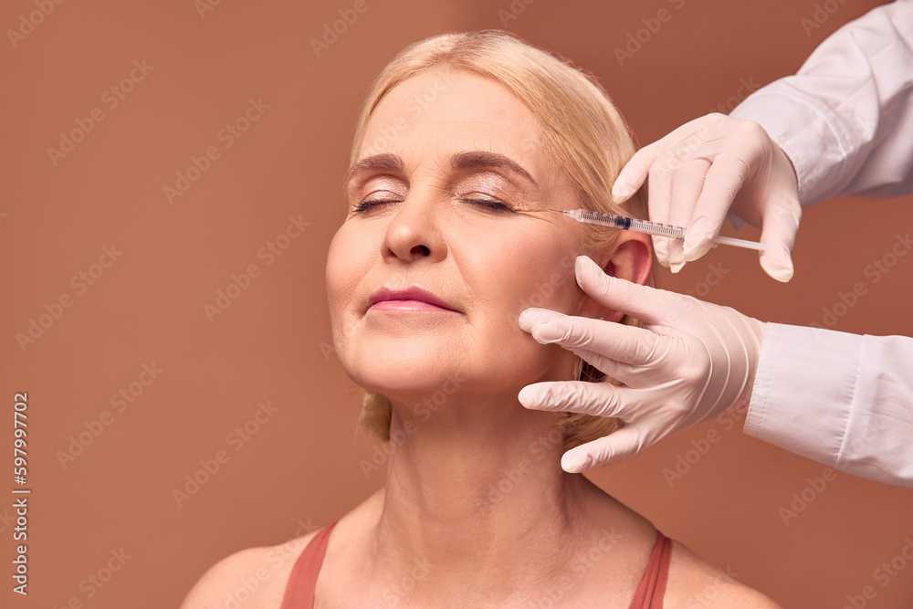 Smoothing out wrinkles.Portrait of an older adult woman with closed eyes  doing a beauty injection. Hands in white gloves hold a syringe.Face lift.  Stock Photo