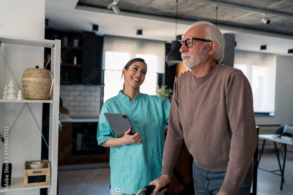Young smiling physiotherapist showing empathy and support to her elderly male patient with disability while he's walking with walking frame. Physiotherapist with senior man. Young smiling physiotherapist showing empathy and support to her elderly male patient with disability while he's walking with walking frame. Physiotherapist with senior man.