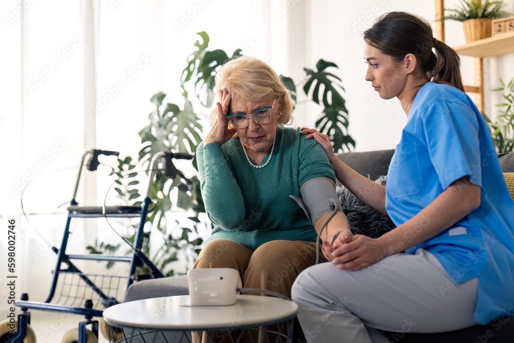 Worried senior woman with hand on her temple looking upset at blood pressure monitor screen concerned about high blood pressure level. Supportive nurse consoling female patient with hypertension Worried senior woman with hand on her temple looking upset at blood pressure monitor screen concerned about high blood pressure level. Supportive nurse consoling female patient with hypertension