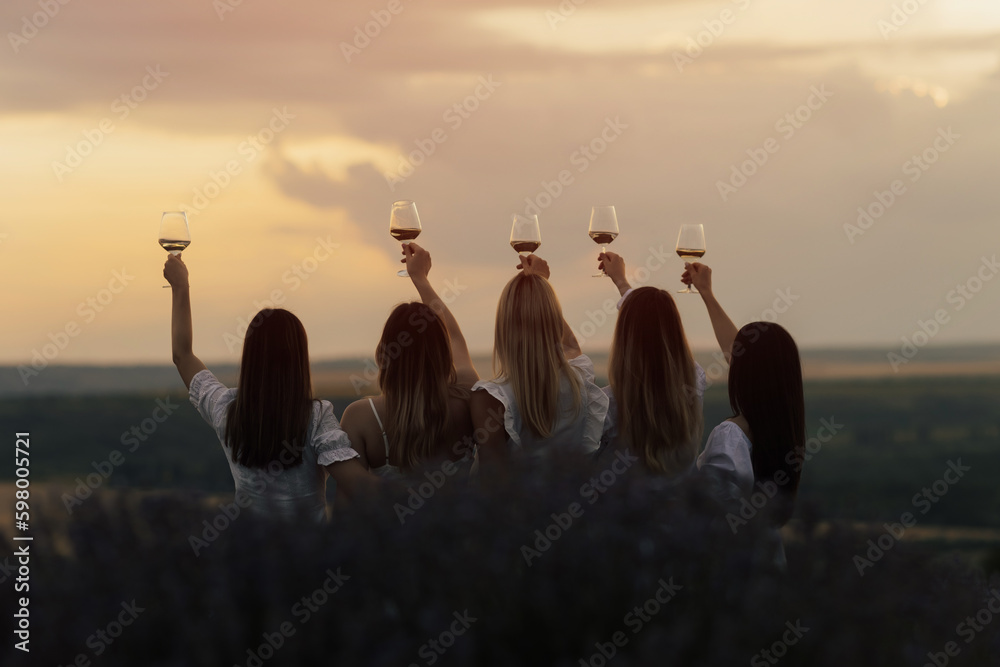 Back view of company of young female friends with hands up having fun, raising glasses with wine and enjoy beautiful sunset at summer picnic in lavender field.