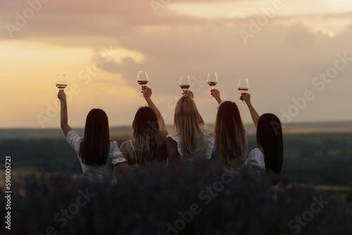 Obraz na płótnie Back view of company of young female friends with hands up having fun, raising glasses with wine and enjoy beautiful sunset at summer picnic in lavender field. w salonie