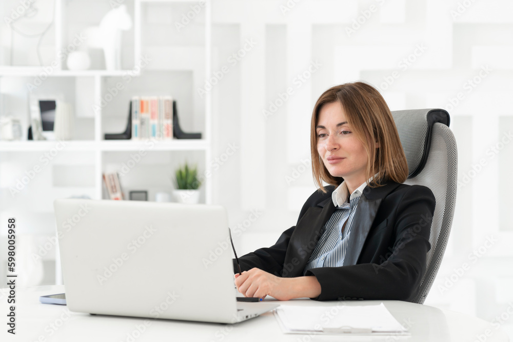 Confident and successful business lady sits at the desk and works on laptop in her modern office Confident and successful business lady sits at the desk and works on laptop in her modern office