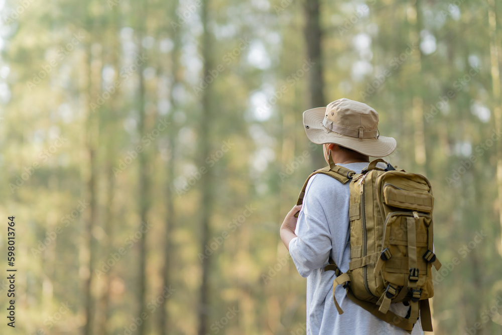 Young Asian man with a backpack and hat hiking in the mountains during the summer season, a traveler walking in the forest. Travel, adventure, and journey concept.