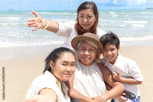 Happy disabled senior elderly woman in wheelchair spending time together with family on summer holiday vacation. Asian grandma, daughter and grandchild boy taking selfie at beautiful tropical beach.