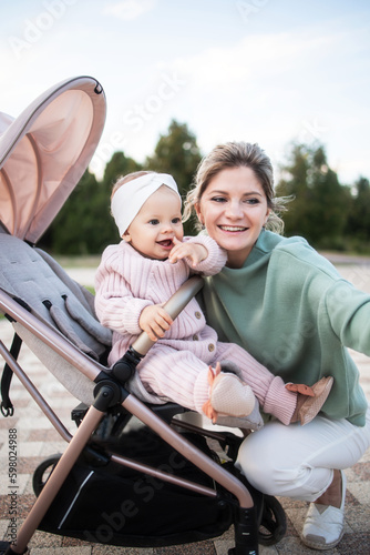Happy positive smiling young mom taking a selfie with her daughter Infant outside while walking in a stroller. Maternity leave. Maternity, Day, Mothers, Parenthood. Stylish woman, baby in stroller