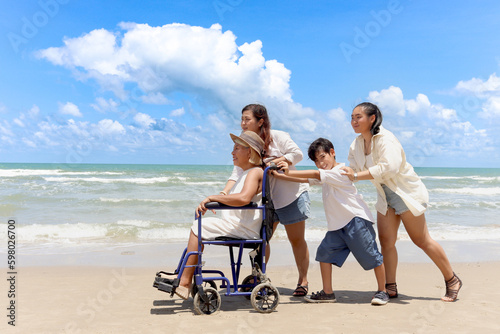 Happy disabled senior elderly woman in wheelchair spending time together with her family on tropical beach. Asian grandma, daughter and grandchild boy resting and relaxing on summer holiday vacation.