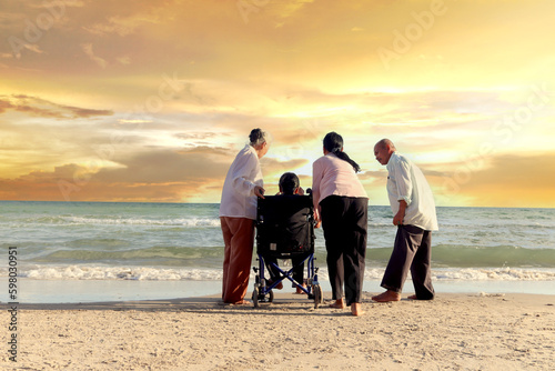 Group of four cheerful elderly old senior have trip outdoor, happy disabled senior elderly woman in wheelchair travel with friends on sunset beach from behind, enjoy together on holiday vacation.