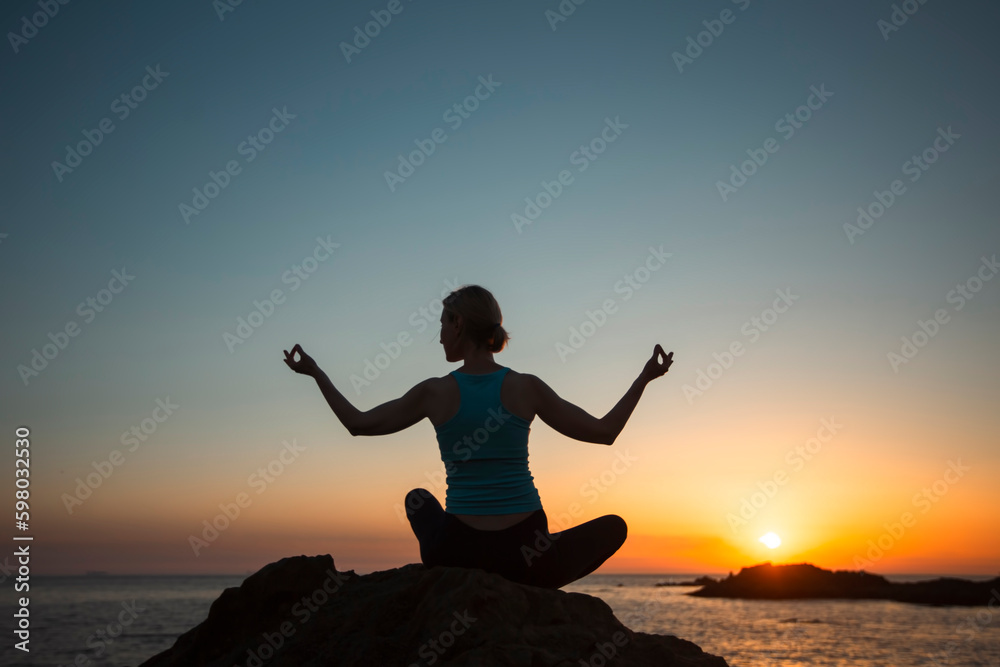 A silhouette of a yoga woman sitting on the ocean shore during sunset.
