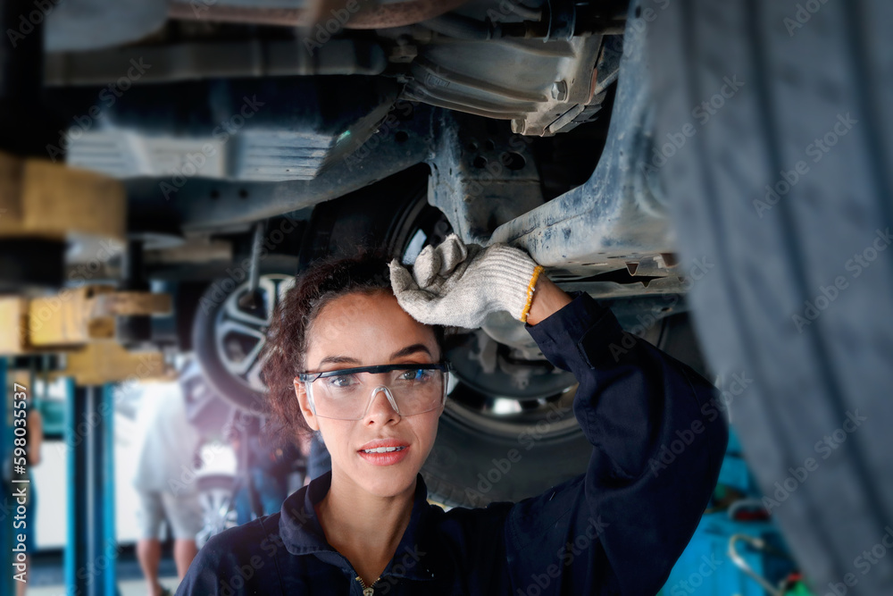 Beautiful female auto mechanic checking wheel tires in garage, car service technician woman repairing customer car at automobile service, inspecting vehicle underbody and suspension engine system.