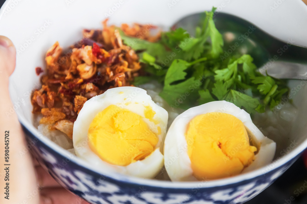 Woman hand holding a bowl of rice congee and boiled eggs with fried garlic, chili and herbs.
