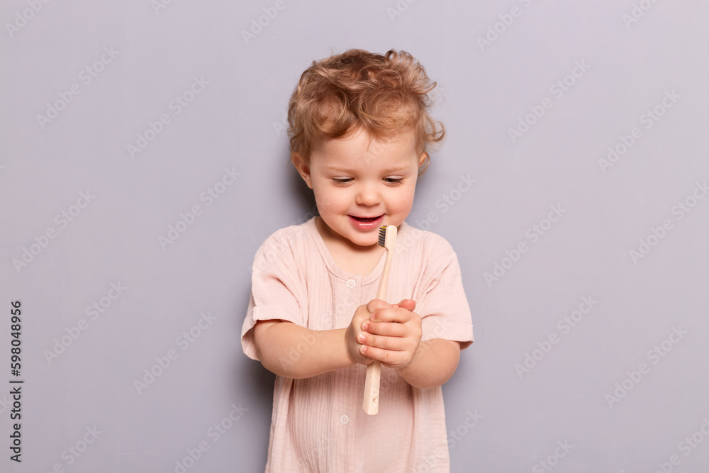 Cute little child baby brushing teeth isolated over gray background holding looking at toothbrush with curious happy face learning taking care of her teeth by herself. Cute little child baby brushing teeth isolated over gray background holding looking at toothbrush with curious happy face learning taking care of her teeth by herself.