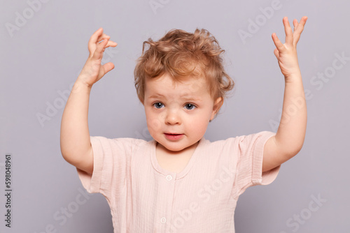 Amazed serious little caucasian toddler girl with blond wavy hair wearing casual shirt standing with raised arms showing how high she will grow posing isolated over gray background
