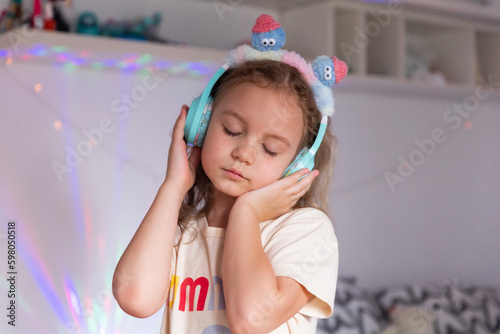 Cute 7 year old girl with headphones listening to music in her room against a background of colored light, portrait. The concept of free time and hobbies of a modern child