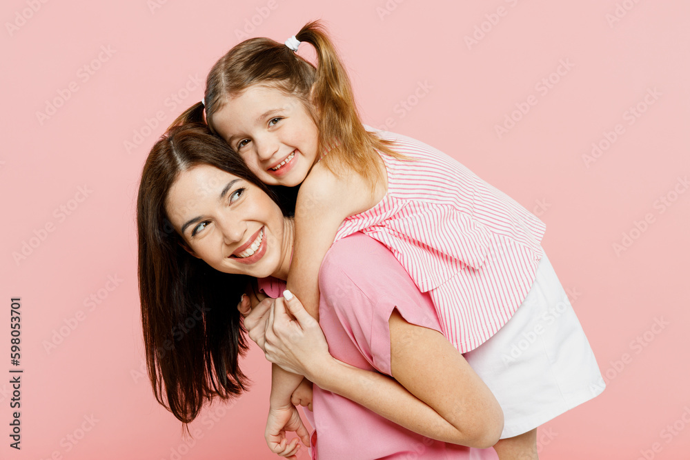 Happy woman wearing casual clothes with child kid girl 6-7 years old. Mother, giving piggyback ride to joyful daughter, sit on back isolated on plain pastel pink background. Family parent day concept. Happy woman wearing casual clothes with child kid girl 6-7 years old. Mother, giving piggyback ride to joyful daughter, sit on back isolated on plain pastel pink background. Family parent day concept.