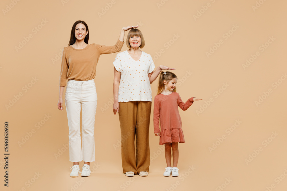 Full body smiling women wearing casual clothes with child kid girl 6-7 years old. Granny mother daughter show height hold hand above head isolated on plain beige background. Family parent day concept.