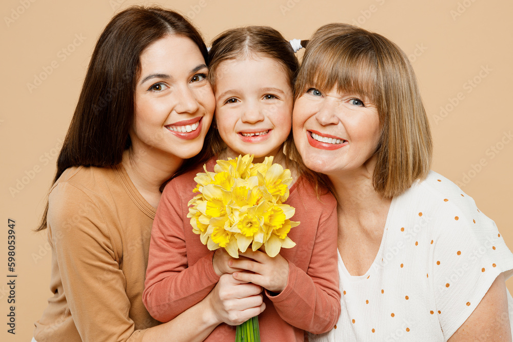 Close up happy women wear casual clothes with child kid girl 6-7 years old. Granny mother daughter gift giving bouquet of daffodil flowers isolated on plain beige background Family parent day concept Close up happy women wear casual clothes with child kid girl 6-7 years old. Granny mother daughter gift giving bouquet of daffodil flowers isolated on plain beige background Family parent day concept