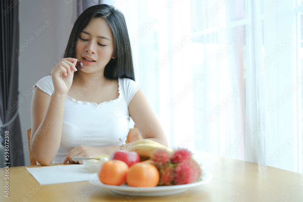 An asian pregnant woman sitting at woodent table beside the window with fruits plate and eating cherry, mum choosing healthy food for baby.