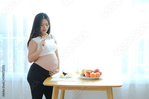 An asian pregnancy woman standing near window with fruits plate on table and eating some of fruit with fork. Mum choosing healthy food for baby.