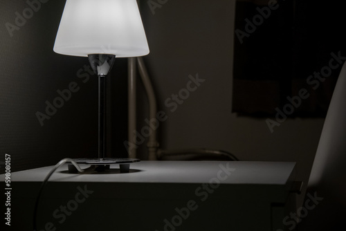 A bedside table and a burning lamp stands on it in a dark room. Hotel number.