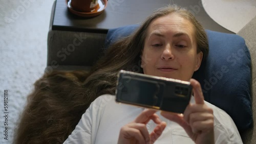 Satisfied happy elderly long haired woman lying with smartphone on sofa photo