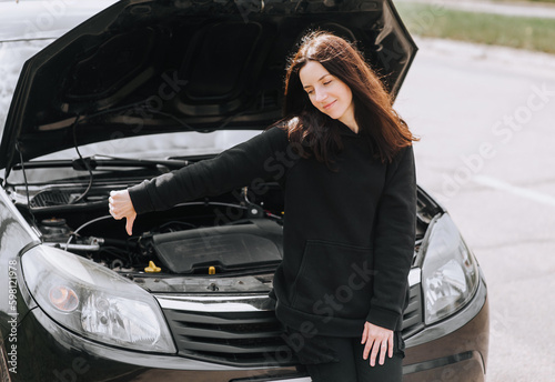 A beautiful young woman stands near a broken black car with an open hood, showing a finger down, dislike gesture. Photography, portrait, breakage concept.