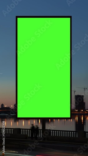 Timelapse green screen billboard on waterfront with night traffic. Vertical lightbox with chroma key on city street. Streetlights reflection on water. Commercial information and Generative AI