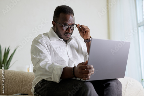 Handsome Black African American Man Working on Laptop Computer while Sitting on a Sofa in Cozy Living Room. Freelancer Working From Home. Browsing Internet, Using Social Networks, Having Fun in Flat.