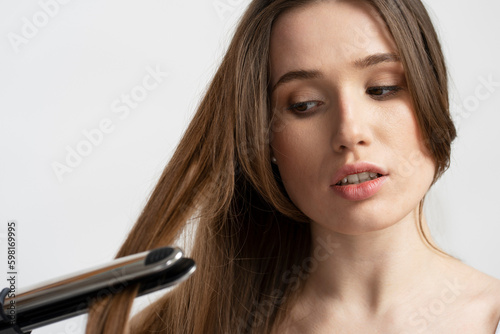 Closeup portrait of beautiful woman using hair straightener doing styling, isolated on white background. Beauty, morning routine, hair care concept