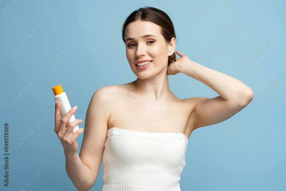 Portrait of young beautiful woman holding bottle with hair shampoo or shower gel looking at camera isolated on blue background. Happy female prepare for shower. Morning routine, body care concept Portrait of young beautiful woman holding bottle with hair shampoo or shower gel looking at camera isolated on blue background. Happy female prepare for shower. Morning routine, body care concept
