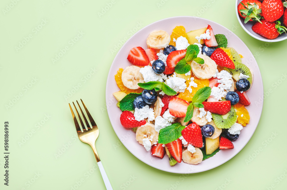 Summer fruit and berry salad with strawberries, blueberries, banana, soft cheese and mint leaves, green background, top view